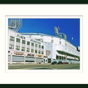 May include: An exterior view of Tiger Stadium, a white building with the words "Tiger Stadium" in blue letters. The stadium is surrounded by a blue sky and a few trees.