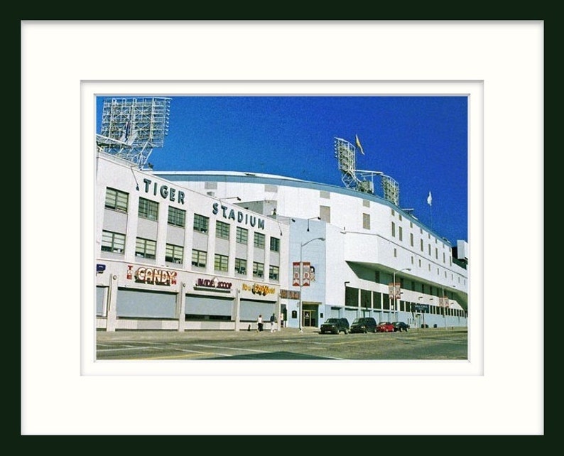 May include: An exterior view of Tiger Stadium, a white building with the words "Tiger Stadium" in blue letters. The stadium is surrounded by a blue sky and a few trees.