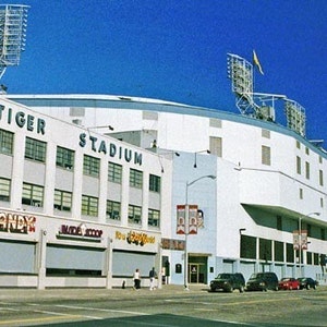 May include: An exterior view of Tiger Stadium, a white building with blue trim and the words "Tiger Stadium" in blue lettering. The stadium is surrounded by a blue sky and a few cars are parked in front of it.