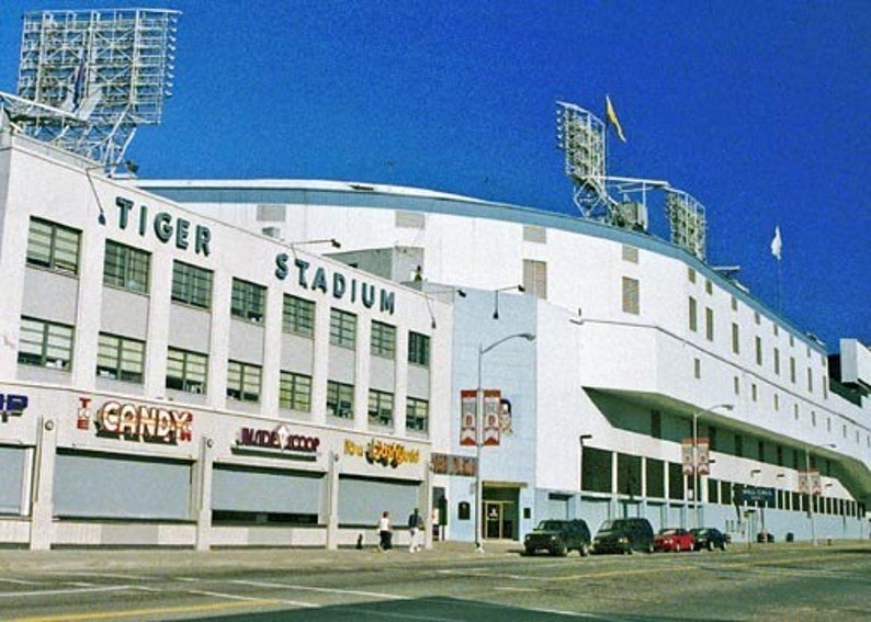May include: An exterior view of Tiger Stadium, a white building with blue trim and the words "Tiger Stadium" in blue lettering. The stadium is surrounded by a blue sky and a few cars are parked in front of it.