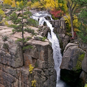 May include: A scenic waterfall cascades over rocky cliffs, surrounded by lush greenery and trees with vibrant yellow and red foliage. The water plunges into a deep gorge, creating a picturesque natural landscape.