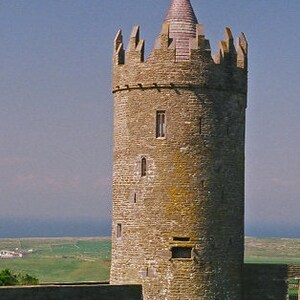 May include: A tall, stone castle tower with a conical roof and crenellations. The tower is cylindrical and has small rectangular windows. The roof is a light pink color, and the sky is a clear blue. The tower is surrounded by green grass.