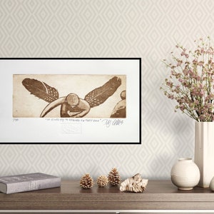 Op de afbeelding: Een ingelijst sepia-gekleurd drukwerk van een tekening van een engel met vleugels van veren. De tekst onder de tekening luidt "Ik zie elke dag de wonderen in een klein zaadje." De handtekening van de kunstenaar is "My Collins".