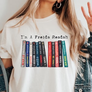 May include: A white t-shirt featuring the text "I'm A Freida Readah" above a graphic illustration of thirteen colourful book spines. Each book spine displays the title and author name, Freida McFadden, in a vertical orientation with varied colours.