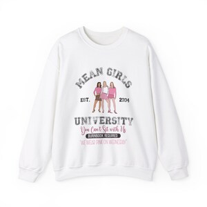 Pu&ograve; includere: Felpa bianca con una stampa grafica di tre ragazze in abiti rosa. Il testo recita "Mean Girls University Est. 2004 You Can't Sit with Us Burnbook Required 'We Wear Pink on Wednesday'"