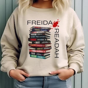 May include: A beige crewneck sweatshirt featuring a graphic of a vertical stack of books by Freida McFadden. The text reads FREIDA READAH with a red paint splatter design. The books have various coloured spines including red, black, and teal.