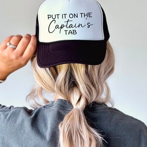 May include: A black and white trucker hat with the text "Put it on the Captain's Tab" in black script font.