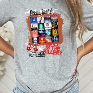 May include: A gray t-shirt featuring a graphic design with the text "Freida Readah" and a collection of book covers. The design includes the text "Who is the Killer?" and "Don't talk to me while I'm reading."