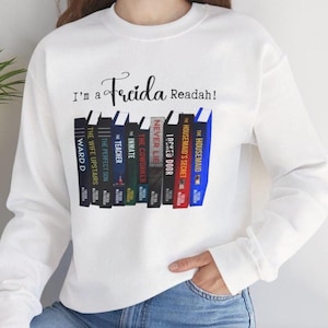 May include: White sweatshirt featuring the phrase "I'm a Frida Readah!" and a graphic of book spines. Titles include "The Housemaid," "The Housemaid's Secret," and "Ward D."