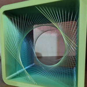 May include: A green, square-shaped, 3D printed object with a swirling, multi-colored, string-like design inside. The design creates a visual illusion of a tunnel or vortex.