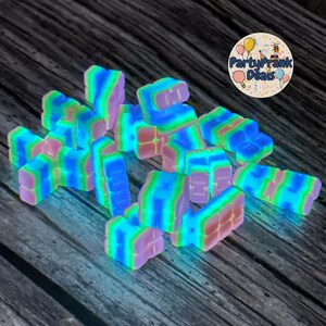 K&ouml;nnte beinhalten: Eine Ansammlung leuchtender, farbenfroher, rechteckiger Bl&ouml;cke in Rosa-, Blau-, Gr&uuml;n- und Lilat&ouml;nen. Die Bl&ouml;cke sind auf einer dunklen Holzoberfl&auml;che verstreut. Ein kreisf&ouml;rmiges Logo mit dem Text "PartyPrank Deals" ist zu sehen.