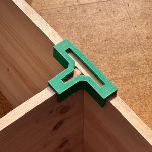 May include: A green corner clamp sits on the corner of two wooden boards. The clamp is made of a solid green material and has a unique shape with a cutout design. The wood appears to be unfinished.