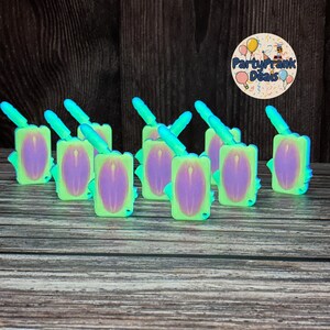 Può includere: Una collezione di bomboniere verde neon e viola. Ogni bomboniera ha un corpo rettangolare con un ovale viola al centro, un manico blu e pinne blu. Le bomboniere sono disposte su una superficie di legno.