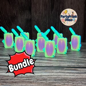 Può includere: Un pacchetto di giocattoli novità. Ogni giocattolo è un rettangolo verde chiaro con un ovale viola e un'antenna blu. La parola "Bundle" è in un fumetto rosso. Lo sfondo è in legno scuro.