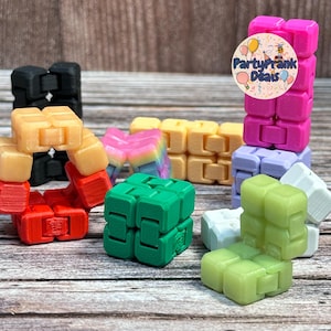 May include: A selection of colourful fidget cubes in shades of black, red, yellow, green, pink, purple, and white. The cubes are displayed on a wooden surface, highlighting their modular design and interlocking parts. The image includes a logo that says "PartyPrank Deals".