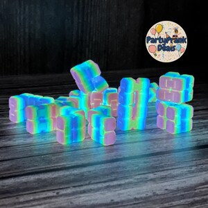 K&ouml;nnte beinhalten: Eine Ansammlung farbenfroher, leuchtender Spielzeugbl&ouml;cke. Die Bl&ouml;cke haben einen Regenbogeneffekt aus Rosa, Blau, Gr&uuml;n und Gelb. Die Bl&ouml;cke sind in einem Haufen angeordnet, einige sind aufeinander gestapelt. Der Hintergrund ist dunkel.