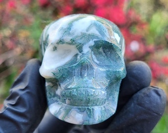 May include: A polished moss agate skull carving featuring a smooth, translucent surface with distinct white and deep green dendritic inclusions. The stone displays natural patterns resembling moss or foliage throughout the entire skull structure.