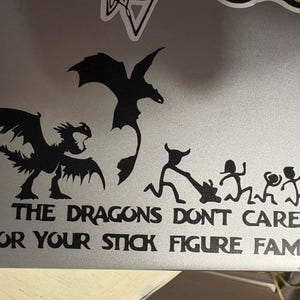 Puede incluir: Pegatina de vinilo negro con siluetas de dragones y figuras de palo. El texto dice "THE DRAGONS DON'T CARE FOR YOUR STICK FIGURE FAMILY". La pegatina está aplicada a un portátil plateado.
