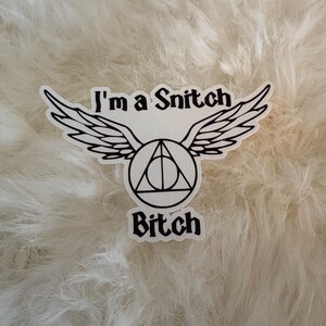 May include: A white sticker with black text and a graphic of wings and a triangle. The text reads "I'm a Snitch Bitch". The sticker is on a white, fluffy surface.