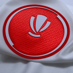 May include: White sports fabric with a red circular emblem. The emblem has a white design inside a red circle, with a white border. The fabric has a textured, breathable weave.