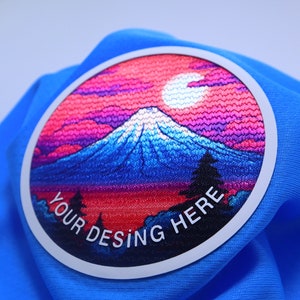 K&ouml;nnte beinhalten: Ein runder gestickter Aufn&auml;her mit einer Bergkette, einem Halbmond und einem rosa und violetten Himmel. Der Text "YOUR DESING HERE" ist um den Rand des Aufn&auml;hers gedruckt.