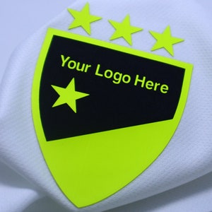 Op de afbeelding: Een close-up van witte stof met een neon groen en zwart embleem. Het embleem is schildvormig met drie sterren aan de bovenkant en een enkele ster aan de linkerkant. De tekst "Your Logo Here" is op het embleem gedrukt.