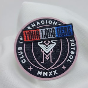 May include: A black and pink round patch with the text "CLUB INTERNACIONAL FÚTBOL" around the edge. The centre of the patch features a white stylised bird design with the text "MMXX" below. A red, white and blue rectangular patch with the text "YOUR LOGO HERE" is on top of the round patch.