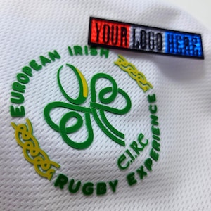 Può includere: Primo piano di tessuto sportivo bianco con un logo a trifoglio verde e la scritta "EUROPEAN IRISH RUGBY EXPERIENCE C.I.R.C." in verde. È visibile anche una piccola toppa rettangolare con la scritta "YOUR LOGO HERE" in rosso, bianco e blu.
