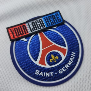 Op de afbeelding: Een wit voetbaltenue met een blauw en rood geborduurd embleem met de Eiffeltoren en de tekst "SAINT-GERMAIN". Een zwart, wit en blauw embleem met de tekst "YOUR LOGO HERE" is bevestigd aan het tenue.