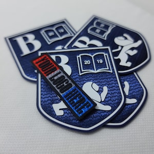 May include: Blue and white embroidered patches with the letter "B" and the year "2019" on a book. The patches are layered on top of each other and have a textured surface. A black and red label with the text "YOUR LOGO HERE" is placed on top of the patches.