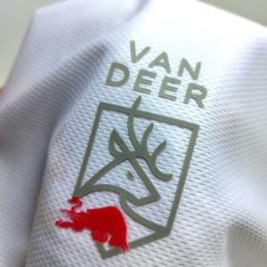 May include: Close-up of a white athletic shirt with a gray logo that reads "VAN DEER". The logo features a deer head inside a gray square with a red bull graphic.