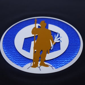 May include: A blue and white circular patch with a gold silhouette of a person holding a spear and a branch. The patch has a textured surface.