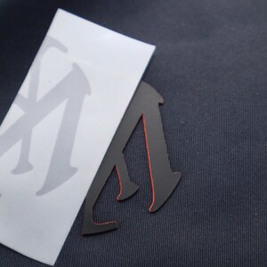 May include: A black vinyl sticker with a red outline in the shape of a person walking. The sticker is on top of a white sheet of paper with a gray letter "M" printed on it.