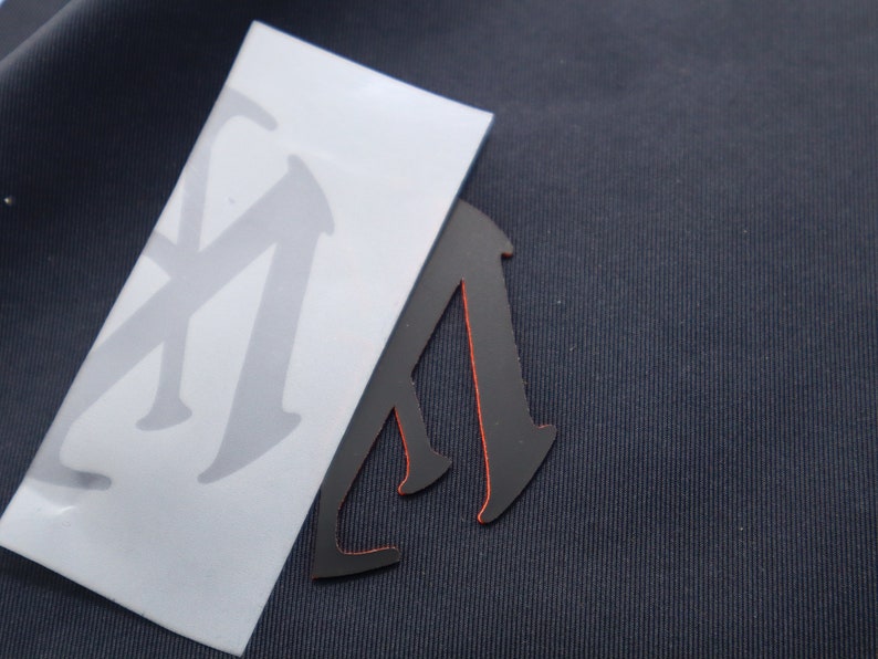 May include: A black vinyl sticker with a red outline in the shape of a person walking. The sticker is on top of a white sheet of paper with a gray letter "M" printed on it.