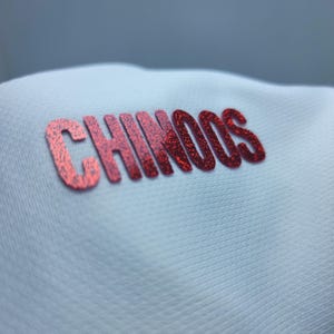 May include: Close-up of a white fabric with the word "CHINOOS" printed in red glitter.