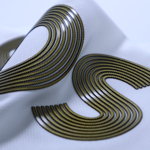 Op de afbeelding: Witte stof met een geborduurd ontwerp in goud en zwart van overlappende gebogen lijnen.