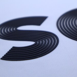 May include: Close-up of a white fabric with the letters "SO" in black, raised and outlined with multiple lines.