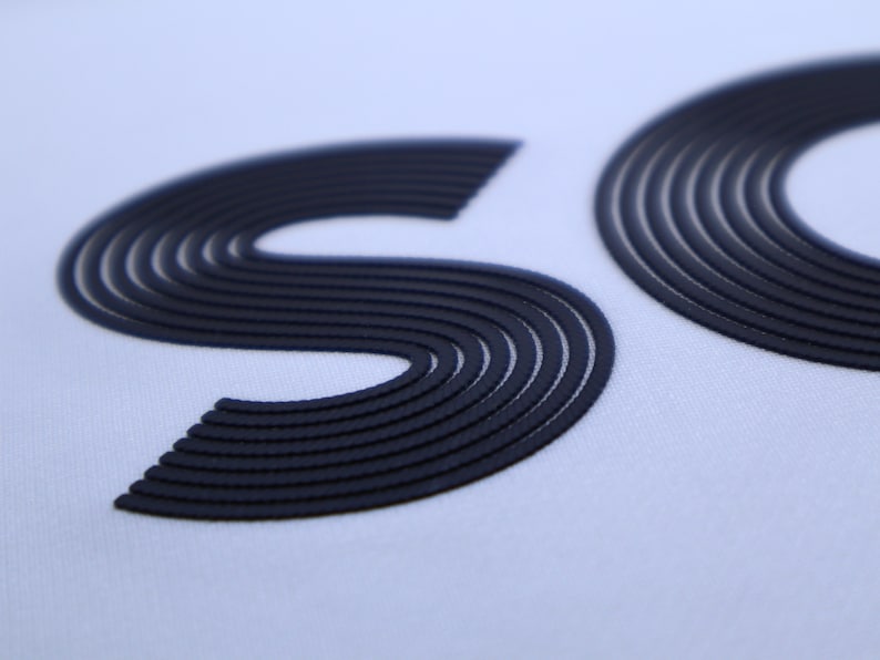 May include: Close-up of a white fabric with the letters "SO" in black, raised and outlined with multiple lines.