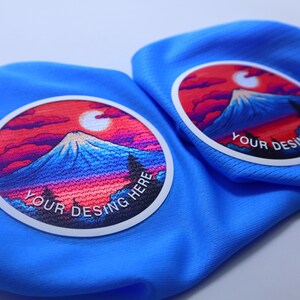 K&ouml;nnte beinhalten: Zwei blaue Stoffpatches mit wei&szlig;en R&auml;ndern und bunten Bergszenen. Der Text "YOUR DESING HERE" ist auf jedem Patch gedruckt.