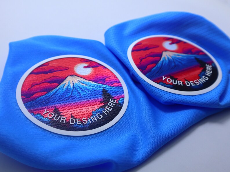K&ouml;nnte beinhalten: Zwei blaue Stoffpatches mit wei&szlig;en R&auml;ndern und bunten Bergszenen. Der Text "YOUR DESING HERE" ist auf jedem Patch gedruckt.