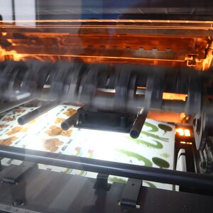 May include: A close-up of a printing press with a sheet of paper with a cartoon bear design moving through the machine.