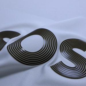 Pode incluir: Um tecido branco com as letras "SOS" impressas em c&iacute;rculos conc&ecirc;ntricos pretos e dourados.