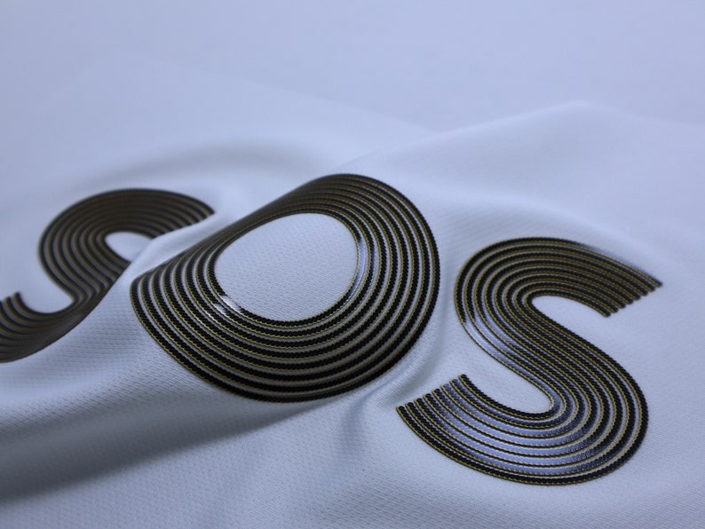 Pode incluir: Um tecido branco com as letras "SOS" impressas em c&iacute;rculos conc&ecirc;ntricos pretos e dourados.