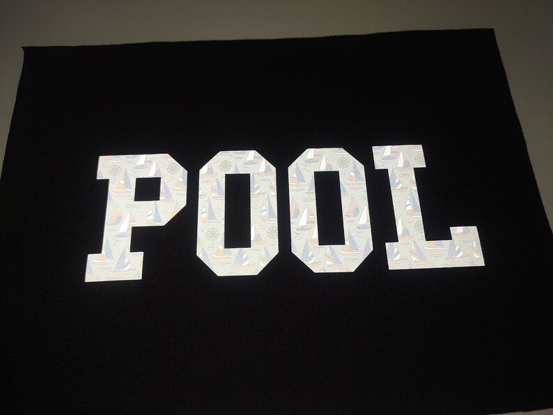 May include: White reflective fabric patch with the word "POOL" in large, bold letters. The letters are filled with a pattern of small sailboats.
