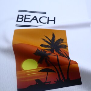 May include: White t-shirt with a graphic print of a sunset with palm trees and the word "BEACH" in black text.