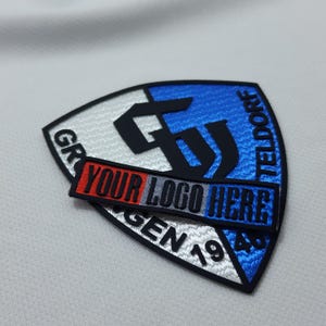Può includere: Una toppa ricamata in nero, bianco e blu con il testo "GR" e "TELDORF" sui lati. Il centro della toppa presenta una "G" e una "J" nere su uno sfondo bianco e blu. Un nastro rosso, bianco e nero è attaccato alla parte inferiore della toppa con il testo "YOUR LOGO HERE".
