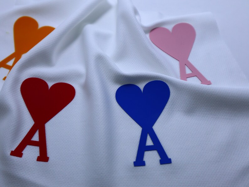 May include: Four heart-shaped patches with the letter 'A' in the center. The hearts are orange, red, blue, and pink. The patches are on a white fabric background.