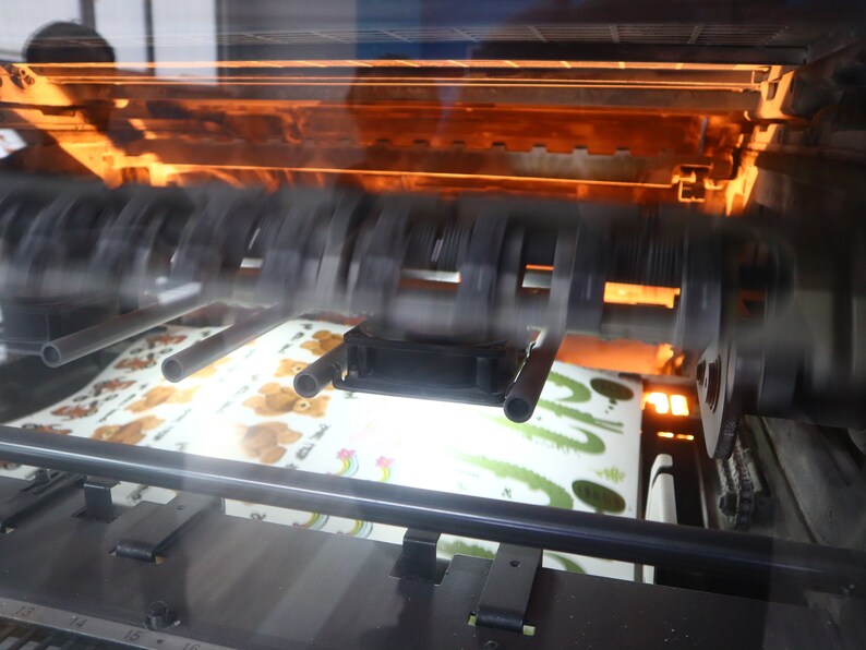 May include: A close-up of a printing press with a sheet of paper with a cartoon bear design moving through the machine.