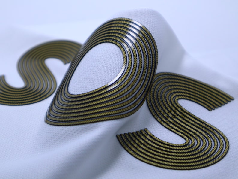 Op de afbeelding: Close-up van een witte stof met een gouden en zwarte "S"-vorm. De stof is getextureerd en de "S" is gemaakt van meerdere overlappende lijnen.