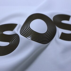 Pode incluir: Um tecido branco com a palavra "SOS" impressa em preto e dourado. As letras s&atilde;o feitas de m&uacute;ltiplos c&iacute;rculos conc&ecirc;ntricos.